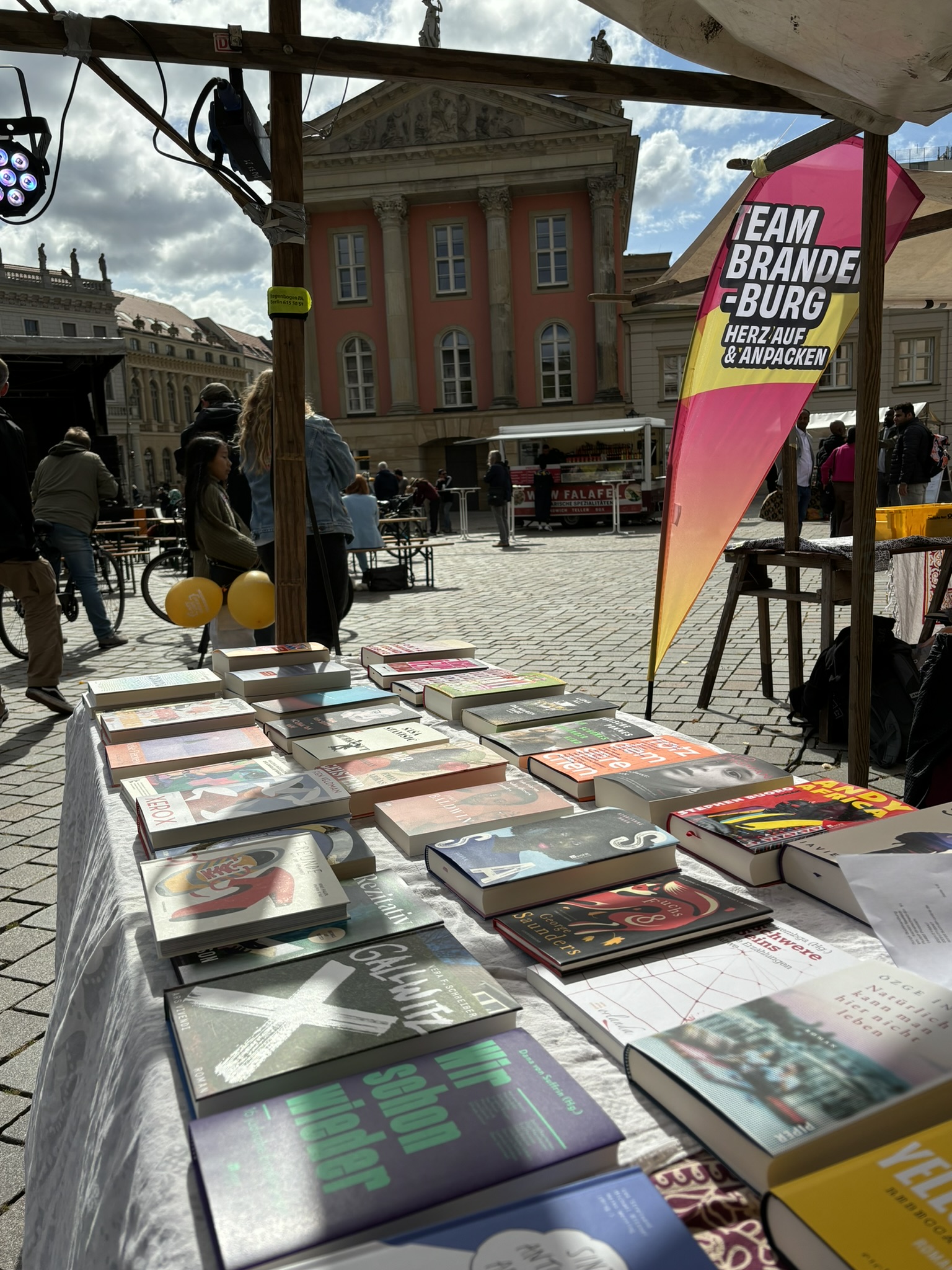 Bücher auf einem Marktstand im Freien vor historischen Gebäuden mit Menschen im Hintergrund und einem pink-gelben Banner mit der Aufschrift "TEAM BRANDENBURG – HERZ AUF & ANPACKEN"